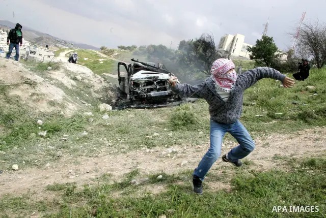 Masked Palestinian youths hurl stones toward Israeli soldiers near a burnt Israeli army vehicle on April 22, 2011 after it flipped over and was set alight by Palestinian youths in the flashpoint neighbourhood of Issawiya...