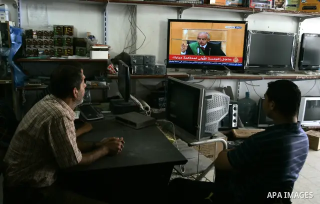 Palestinian men watch the video image taken from Egyptian State Television at a shop in Rafah, southern Gaza Strip, where shows 83-year-old Hosni Mubarak laying on a hospital bed inside a cage of mesh and iron bars as he...