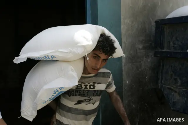 A Palestinian youth carries a bag with wheat flour on his shoulders as others receive food aids from the United Nations Relief and Works Agency (UNRWA) at the Rafah refugee camp, in the southern Gaza Strip on August 15,...