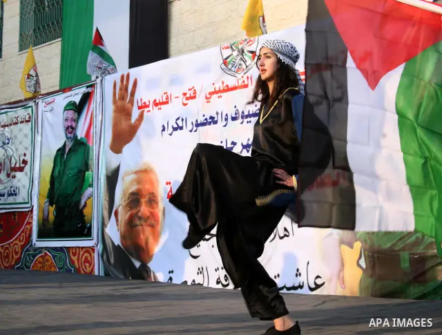 Palestinian folkloric dancers perform in front of a poster showing the late Palestinian leader Yasser Arafat during a celebration to to mark the 47th anniversary of the Fatah political movement. in the West Bank city of...