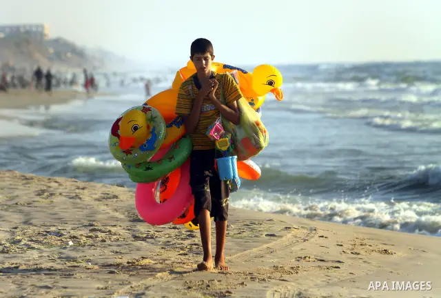 A Palestinian child sells toys as others enjoy swimming in the sea on Gaza's beach on 27 June 2012 in Gaza. Photo by Majdi Fathi