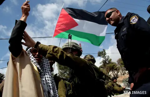 A Palestinian man in traditional garb holds the national flag as he points to an Israeli soldiers during a protest near an Israeli-occupied Palestinian home, located close to the Kiryat Arbaa Jewish settlment, as Palesti...