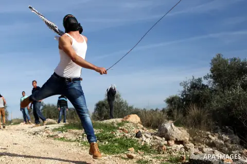 A Palestinian protester uses a slingshot to throw a stone towards Israeli security forces during clashes following a demonstration against Israel's controversial barrier, in the outskirts of the village of Bilin, near th...