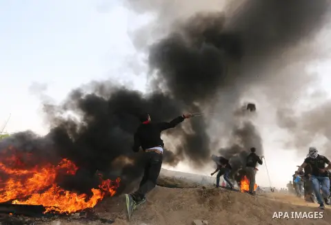 A Palestinian protester uses a slingshot to hurl stones as others burn tires to protect themselves from shots of Israeli soldiers during clashes at the Israel-Gaza border in a tent city protest demanding the right to ret...